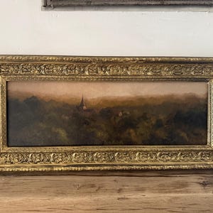 Peut inclure: Une peinture à l'huile paysagère rectangulaire représentant une flèche d'église lointaine s'élevant au-dessus d'une forêt dense et vert foncé sous un ciel brumeux et atténué. L'œuvre est encadrée par un large cadre doré orné de volutes.