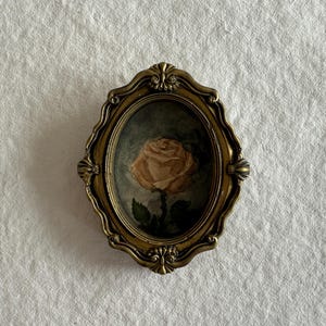 Peut inclure: Un cadre ovale doré de style ancien avec un design orné. À l'intérieur, une peinture d'une rose unique de couleur claire avec des feuilles vertes sur un fond discret. Le cadre est un objet décoratif.