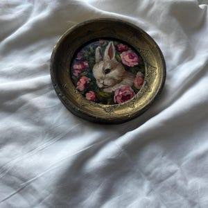 May include: A small, round, gold-colored frame with a painting of a white rabbit surrounded by pink flowers. The frame is on a white surface.