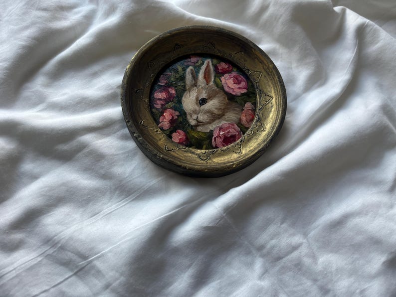 May include: A small, round, gold-colored frame with a painting of a white rabbit surrounded by pink flowers. The frame is on a white surface.