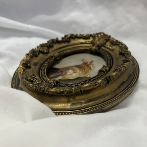 Puede incluir: Un peque&ntilde;o marco de fotos ovalado con acabado dorado. El marco tiene un dise&ntilde;o ornamentado detallado y contiene una pintura de un gato.