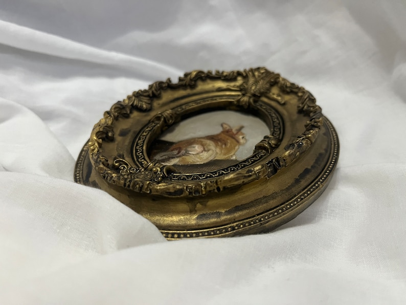 Puede incluir: Un peque&ntilde;o marco de fotos ovalado con acabado dorado. El marco tiene un dise&ntilde;o ornamentado detallado y contiene una pintura de un gato.