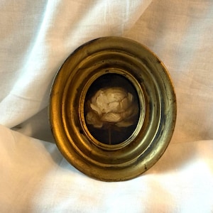Peut inclure: Cadre ovale doré antique avec une peinture d'une fleur blanche. La fleur est centrée sur un fond sombre. Le cadre présente un design détaillé et superposé. L'œuvre est posée sur un tissu blanc texturé.