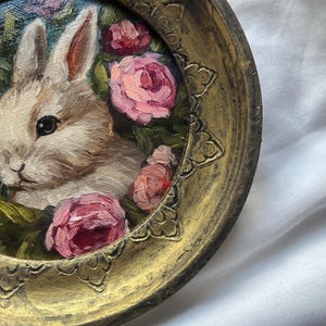 May include: A white rabbit with pink eyes surrounded by pink roses in a gold frame.