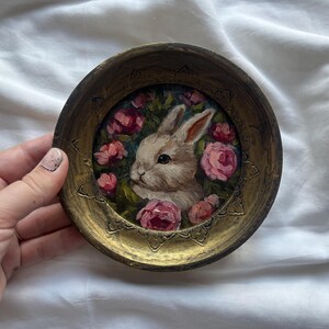 May include: A small, round, gold-colored frame with a painted image of a white rabbit surrounded by pink roses. The rabbit is looking to the left and has a pink nose.