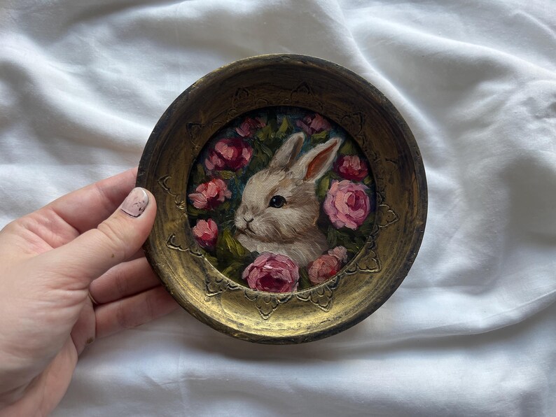 May include: A small, round, gold-colored frame with a painted image of a white rabbit surrounded by pink roses. The rabbit is looking to the left and has a pink nose.