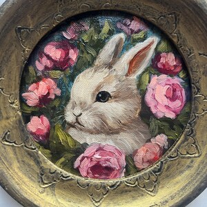 May include: A white rabbit with pink eyes is surrounded by pink roses in a gold painted frame. The rabbit is looking to the left of the image.