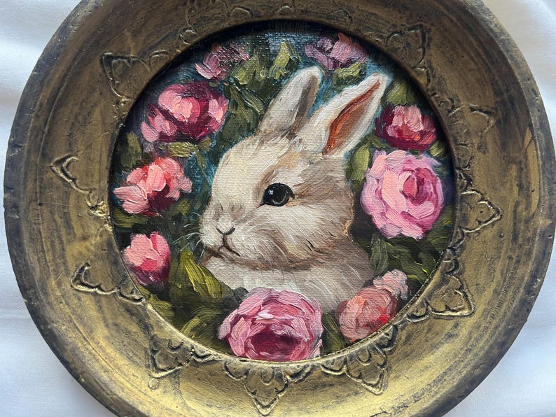 May include: A white rabbit with pink eyes is surrounded by pink roses in a gold painted frame. The rabbit is looking to the left of the image.