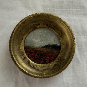 May include: A small, round painting in a gold-colored frame. The artwork depicts a landscape with a red field, dark green hills, and a gray sky. The frame has a decorative, textured edge.