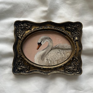 Peut inclure: Peinture ovale d'un cygne dans un cadre doré décoratif. Le cygne est blanc avec un bec noir et se détache sur un fond rose. Le cadre orné présente un design vintage détaillé.