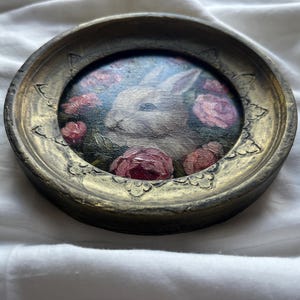 May include: A white rabbit painted in a round frame with pink roses and green leaves. The frame is gold with a decorative design.