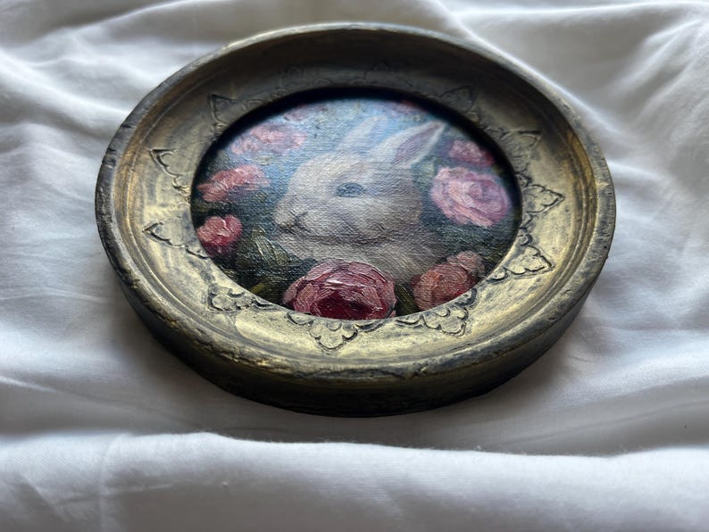 May include: A white rabbit painted in a round frame with pink roses and green leaves. The frame is gold with a decorative design.
