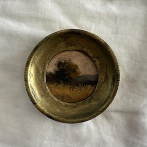 May include: A small, round, antique gold-toned frame with an embossed border. The frame holds a miniature landscape painting featuring a tree, field, and mountains under a pinkish sky. The painting is a vintage piece.