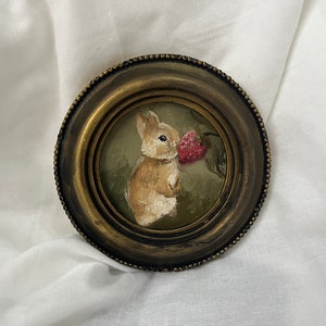 May include: A small, round, antique-style framed painting. The artwork depicts a brown bunny holding a red strawberry against a green background. The frame is a dark, tarnished gold color.