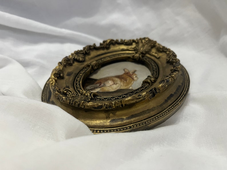 Puede incluir: Un peque&ntilde;o marco de fotos ovalado con acabado dorado. El marco tiene un dise&ntilde;o ornamentado detallado y una peque&ntilde;a pintura de un gato en su interior.