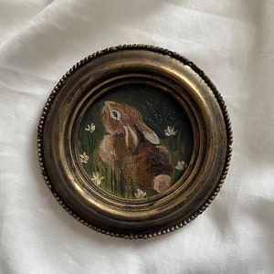 Peut inclure: Un petit cadre rond doré avec une peinture d'un lapin brun regardant vers le haut. Le lapin est entouré de fleurs blanches et de feuillage vert.