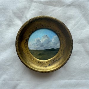 Puede incluir: Una pequeña pintura redonda de un paisaje. La obra de arte representa un cielo azul con nubes blancas sobre un campo verde y una línea oscura del horizonte. La pintura está enmarcada en un marco decorativo de color oro antiguo.