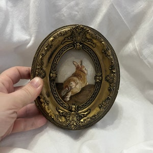 Puede incluir: Un peque&ntilde;o marco dorado adornado con una pintura de un conejo marr&oacute;n y blanco. El conejo est&aacute; sentado en una superficie marr&oacute;n y mirando hacia la derecha.