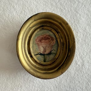Peut inclure: Petite peinture ovale d'une rose rose dans un cadre doré. La rose est placée sur un fond bleu clair. Le cadre présente un design orné. Une œuvre d'art miniature.