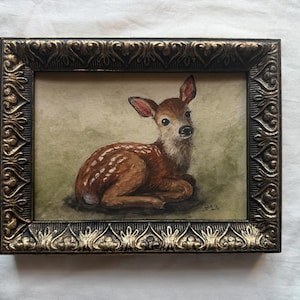 Peut inclure: Une peinture encadrée représentant un jeune faon au repos sur un fond vert doux. Le faon a un pelage brun avec des taches blanches et de grands yeux sombres. Le cadre rectangulaire est sombre avec une finition dorée et un motif en relief.