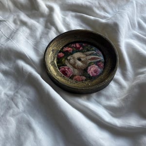 May include: A round, wooden frame with a gold-colored trim encloses a painting of a white rabbit with pink flowers. The rabbit is looking to the left, and the flowers are in bloom.