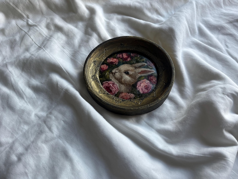 May include: A round, wooden frame with a gold-colored trim encloses a painting of a white rabbit with pink flowers. The rabbit is looking to the left, and the flowers are in bloom.