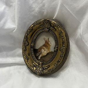 Puede incluir: Una pintura enmarcada de un conejo marr&oacute;n sentado sobre una superficie marr&oacute;n. El marco es dorado y de forma ovalada con detalles ornamentados.