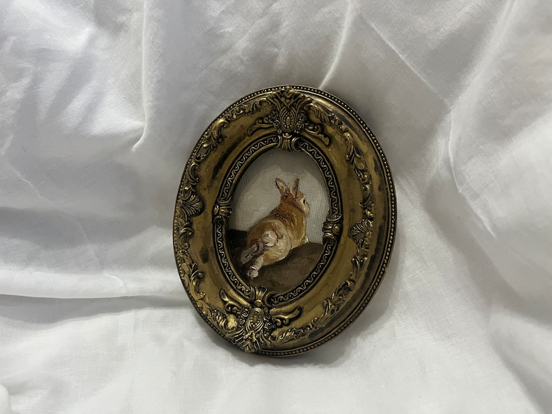 Puede incluir: Una pintura enmarcada de un conejo marr&oacute;n sentado sobre una superficie marr&oacute;n. El marco es dorado y de forma ovalada con detalles ornamentados.