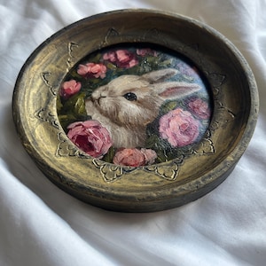 May include: A round, gold-colored frame with a painted image of a white rabbit surrounded by pink roses. The frame has a decorative, ornate design.
