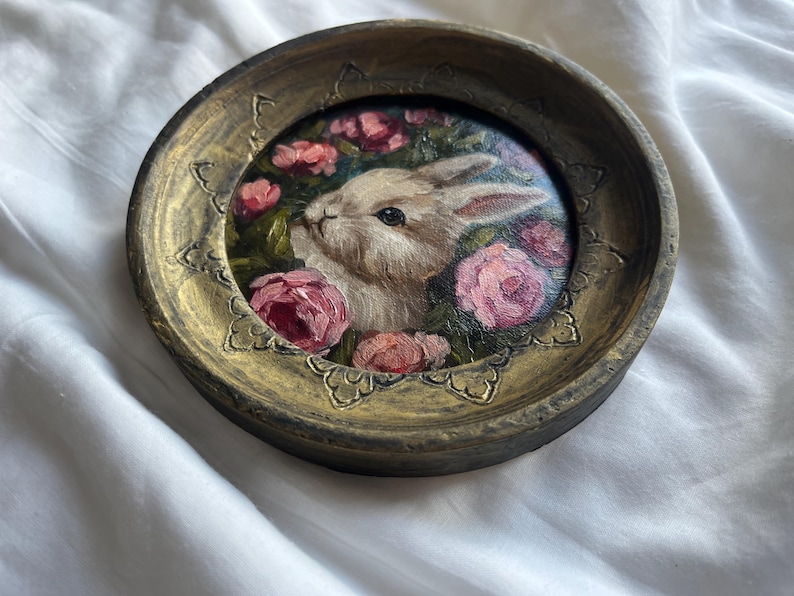 May include: A round, gold-colored frame with a painted image of a white rabbit surrounded by pink roses. The frame has a decorative, ornate design.