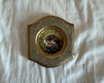 Puede incluir: Una pequeña pintura enmarcada de un erizo. La obra presenta una representación detallada del erizo con púas marrones y blancas, sobre un fondo floral verde y blanco. El marco es ornamentado, con un acabado dorado y plateado.