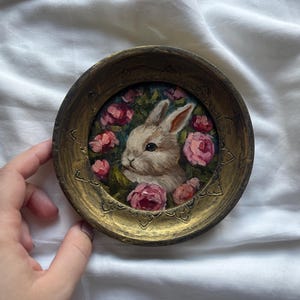 May include: A small, white rabbit painted on a round, gold-colored tray. The rabbit is surrounded by pink flowers and green leaves.