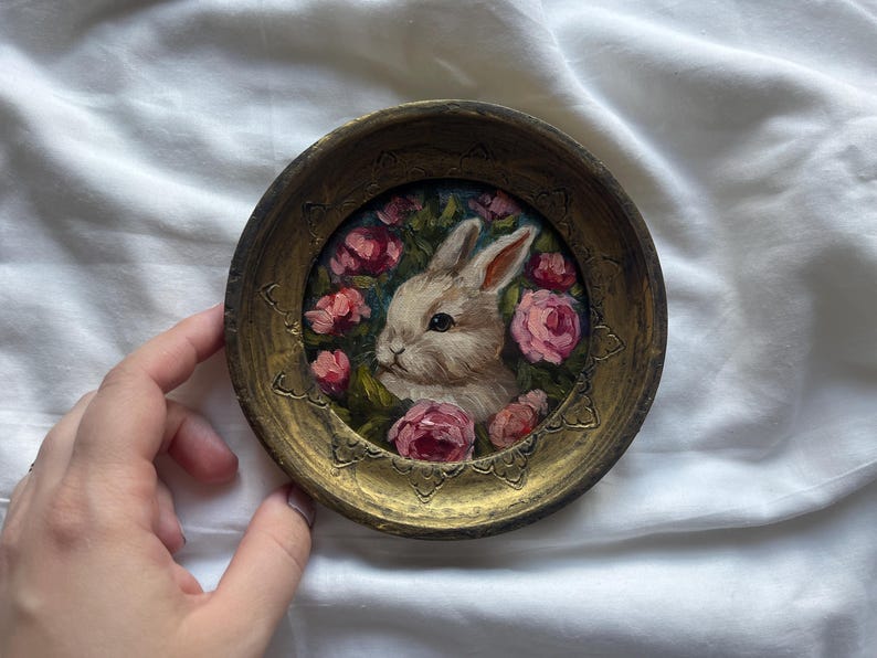 May include: A small, white rabbit painted on a round, gold-colored tray. The rabbit is surrounded by pink flowers and green leaves.