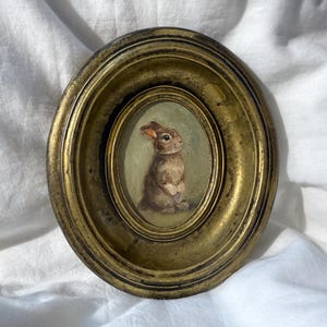 Peut inclure: Un petit tableau ovale d'un lapin brun dans un cadre doré. Le lapin est représenté assis, les oreilles dressées. Le cadre a un aspect ancien. La peinture a une esthétique vintage, avec des couleurs douces et des coups de pinceau.
