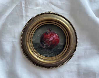 May include: A small, round, gold-framed painting of a red pomegranate on a dark green background.