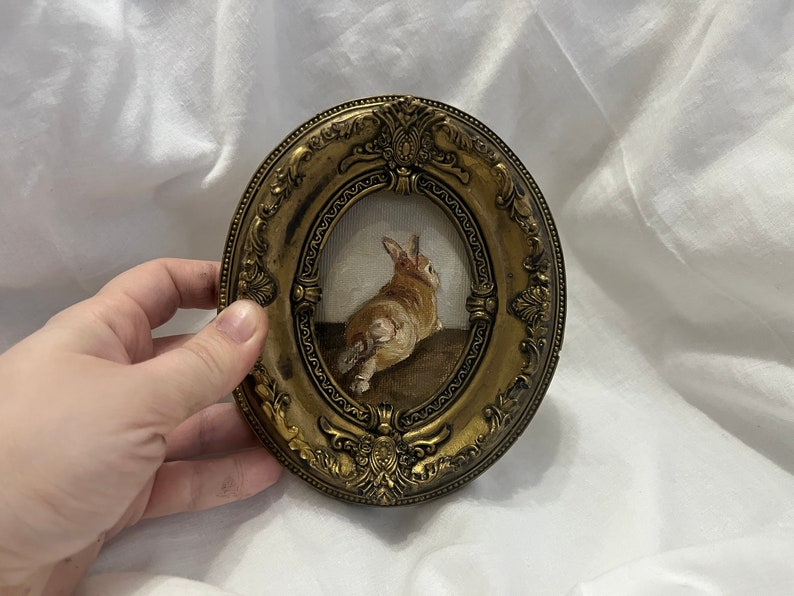 Puede incluir: Un peque&ntilde;o marco dorado adornado con una pintura de un conejo marr&oacute;n y blanco. El conejo est&aacute; sentado en una superficie marr&oacute;n y mirando hacia la derecha.