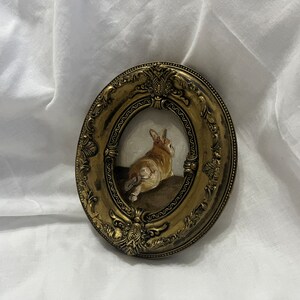 Puede incluir: Un marco peque&ntilde;o, ovalado, de color dorado con detalles ornamentados rodea una pintura de un conejo marr&oacute;n sentado sobre una superficie marr&oacute;n. El conejo est&aacute; de espaldas al espectador.
