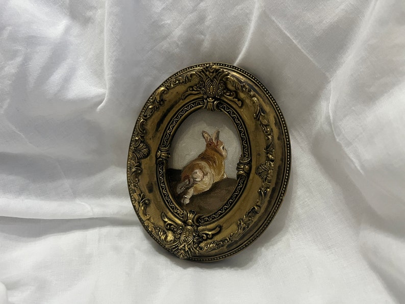 Puede incluir: Un marco peque&ntilde;o, ovalado, de color dorado con detalles ornamentados rodea una pintura de un conejo marr&oacute;n sentado sobre una superficie marr&oacute;n. El conejo est&aacute; de espaldas al espectador.