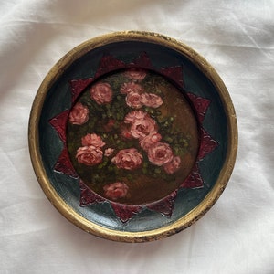 Peut inclure: Un plateau rond en bois avec un bord doré et un motif peint de roses roses sur un fond sombre. Le plateau a un motif géométrique rouge et bleu autour du bord.
