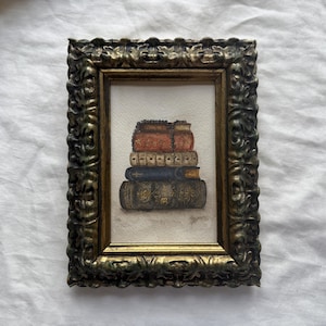 Peut inclure: Une peinture rectangulaire représentant une pile de quatre livres dans un cadre doré ancien, orné et sombre. Les livres sont peints dans des tons de rouge, crème, bleu et gris foncé, avec des détails sur les tranches sur un fond blanc cassé.