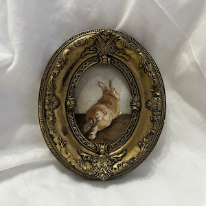 Puede incluir: Una obra de arte de punto de aguja enmarcada que representa un conejo marr&oacute;n y blanco sentado sobre sus patas traseras. El marco es de forma ovalada y tiene un acabado dorado con detalles ornamentados.