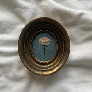 Könnte beinhalten: Ein kleiner, ovaler, goldfarbener Rahmen mit einer weißen Blume auf blauem Hintergrund.