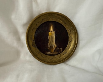 May include: A small, round painting of a lit candle in a gold-colored frame. The candle is off-white with a yellow flame, set against a dark brown background. The frame has a decorative, floral-like pattern.