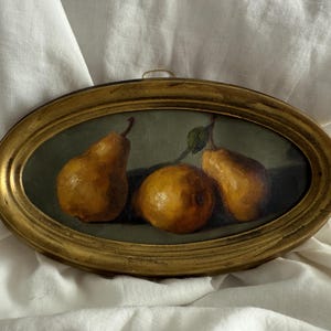 Peut inclure: Peinture de nature morte encadrée ovale de trois poires. Les poires sont brun doré avec une tige et une feuille vertes. Le cadre est doré et le fond est vert. La peinture est posée sur un tissu blanc.