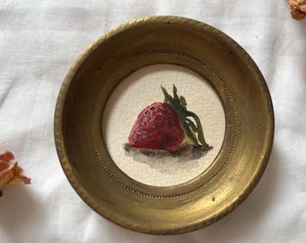 May include: A brass tray with a small round frame containing a watercolor painting of a red strawberry with green leaves.