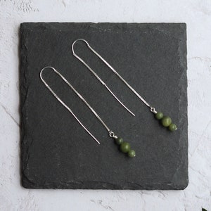 Może przedstawiać: Para srebrnych kolczyków z zielonymi koralikami z jadeitu. Kolczyki są prezentowane na czarnej płytce ze łupka.