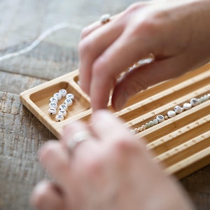 May include: A wooden bead tray with multiple compartments for organizing beads. The tray has a ruler on the side for measuring the length of the string. The tray is filled with white letter beads that spell out the name "ISARA" and other beads in various colors.