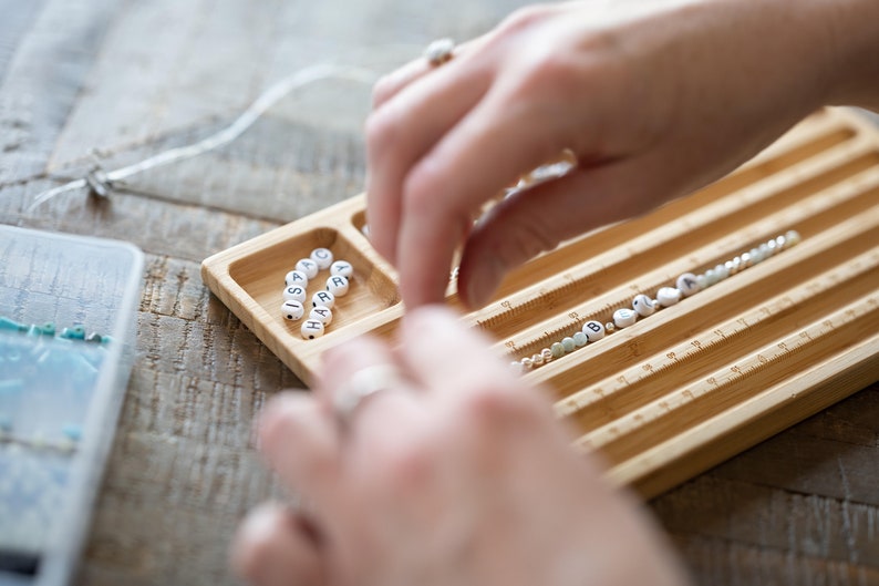 May include: A wooden bead tray with multiple compartments for organizing beads. The tray has a ruler on the side for measuring the length of the string. The tray is filled with white letter beads that spell out the name "ISARA" and other beads in various colors.