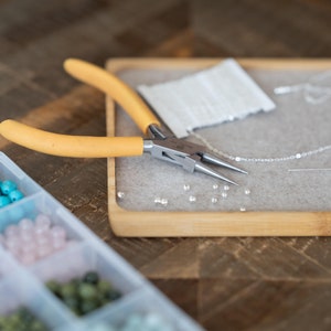 May include: A close-up of a jewelry making kit with a variety of beads, a silver chain, and a pair of yellow-handled pliers.