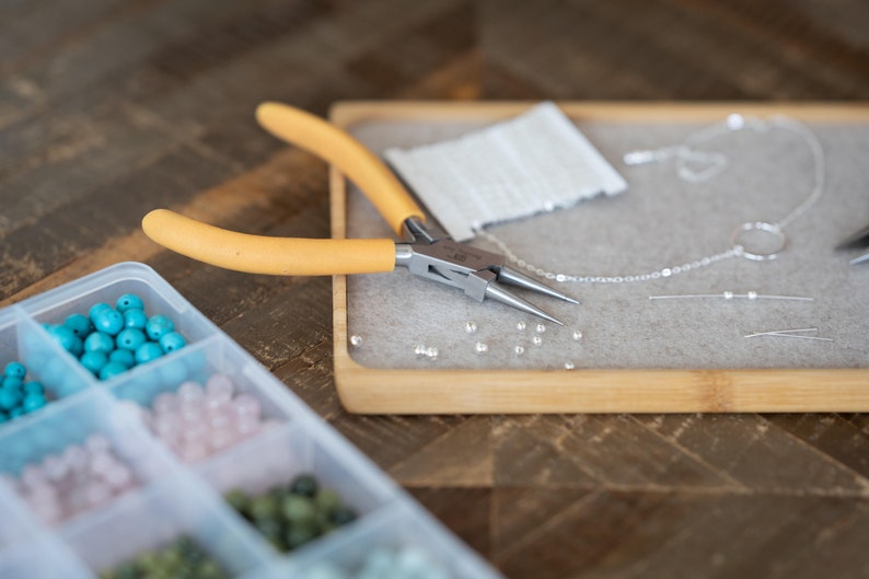 May include: A close-up of a jewelry making kit with a variety of beads, a silver chain, and a pair of yellow-handled pliers.